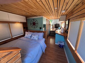 Quarto compacto e bem iluminado, com uma cama, pequeno espaço de cozinha e uma bússola estelar visível na parede.