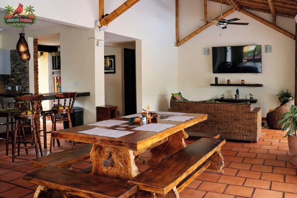 Une table à manger en bois avec des bancs, un coin bar, des meubles en osier, un téléviseur et un ventilateur de plafond dans une pièce dont le sol est recouvert de carreaux.