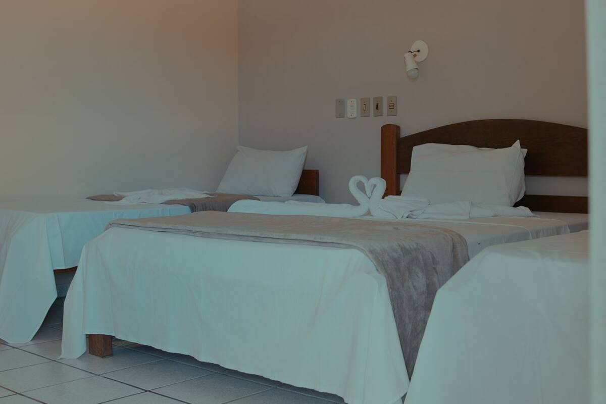 A hotel room with three beds, each with white bedding and a wooden headboard. There are wall sockets and a light fixture above the beds.