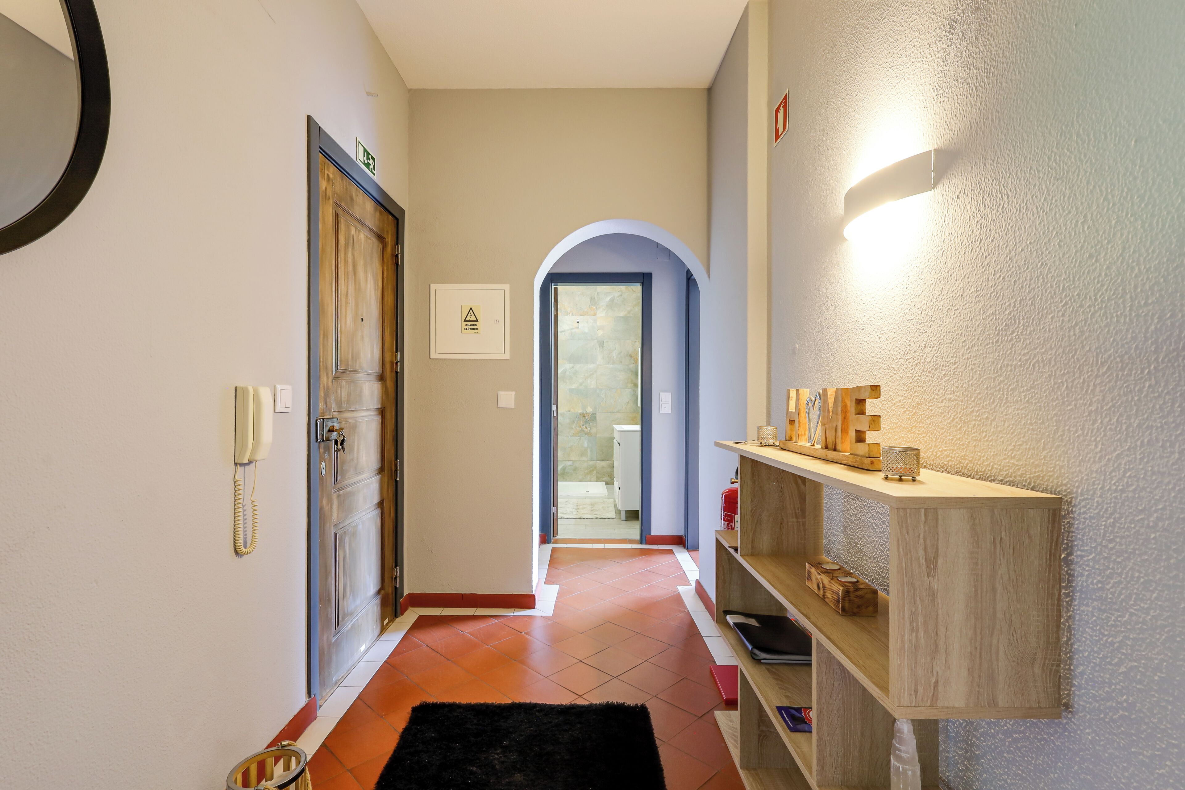 Un couloir avec une porte en bois, un téléphone, une étagère ornée d’objets décoratifs et un sol carrelé.