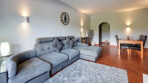 A modern living room with a grey sectional sofa, a dining table with chairs, a wall clock, and a small rug. - Golden Grandfather's Corner (Albufeira)