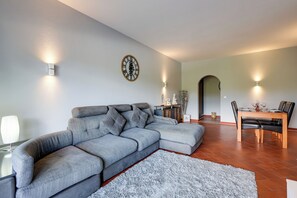 A modern living room with a grey sectional sofa, a dining table with chairs, a wall clock, and a small rug. - Golden Grandfather's Corner (Albufeira)