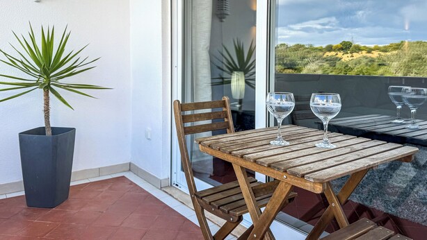 A wooden table with two wine glasses, a potted plant, and a chair on a balcony with a view of the landscape. - Golden Grandfather's Corner (Albufeira)