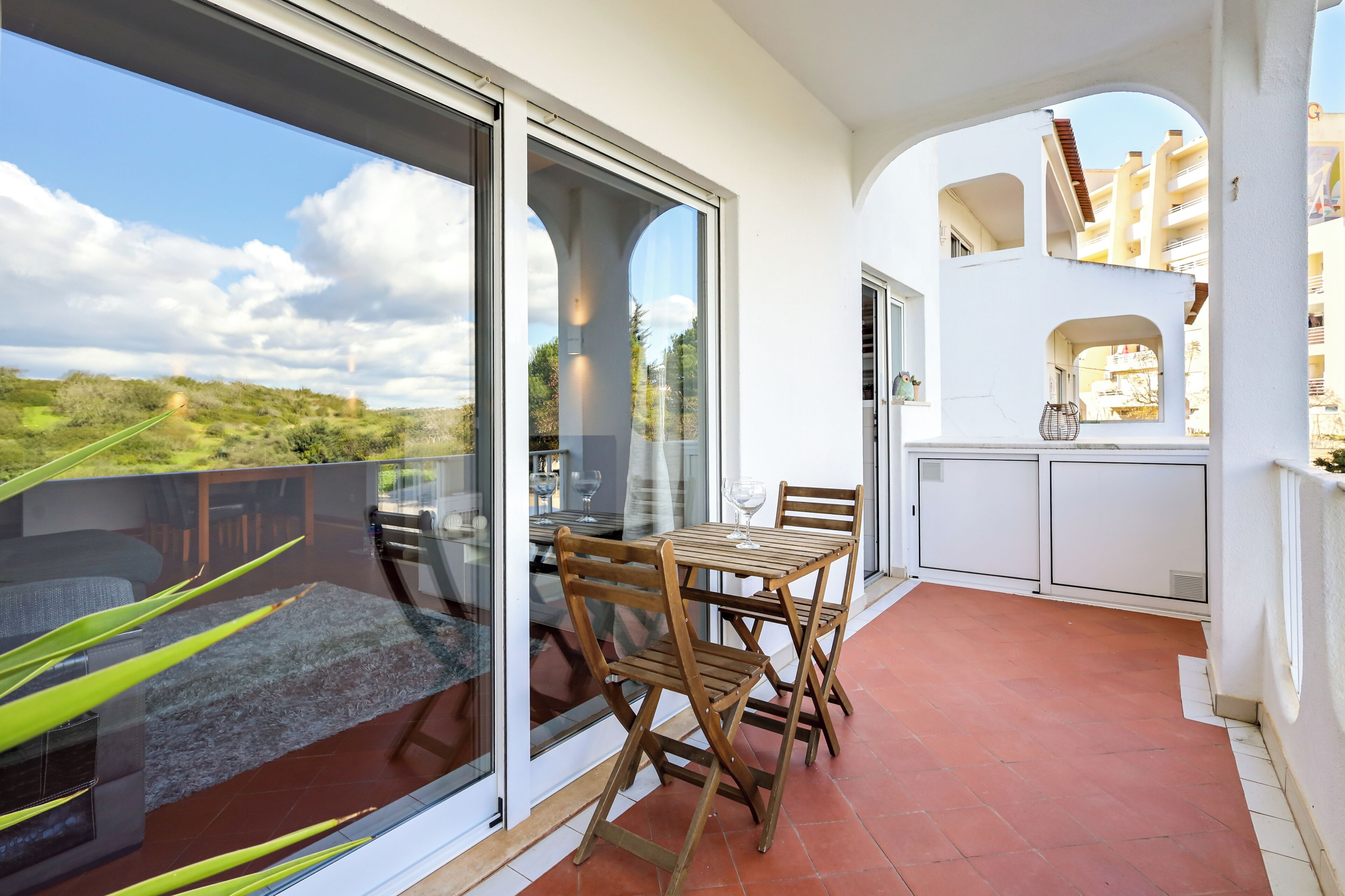 A balcony with a glass door, wooden table, and chairs, overlooking a landscape.