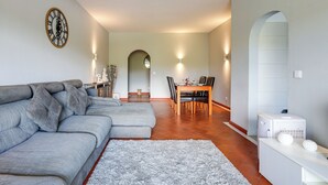 A living room with a sectional sofa, a dining table with chairs, a kitchen area, and a clock on the wall. - Golden Grandfather's Corner (Albufeira)