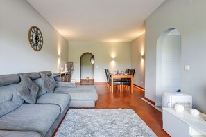 A living room with a sectional sofa, a dining table with chairs, a kitchen area, and a clock on the wall. - Golden Grandfather's Corner (Albufeira)