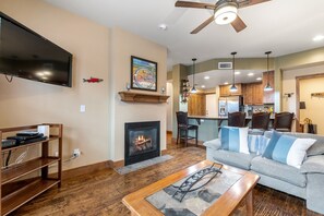 A living room with a fireplace, a television, a ceiling fan, and a kitchen area in the background. - Newly Listed Luxury Whitefish Vacation Rental with Amazing Amenities (Whitefish)