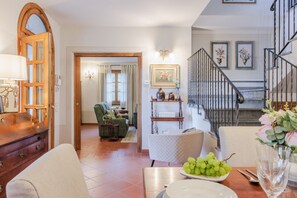 A living room with a sofa, a dining table with a plate of grapes, a staircase, and a chandelier. - Villa Near Lucca With Vineyard Views (Lucca)