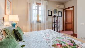 Een slaapkamer met een bed, een nachtkastje met lamp, een raam met gordijnen, een houten deur en een wandplank met decoratieve objecten.