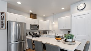 A modern kitchen with white cabinets, stainless steel appliances, a central island with seating, and a clock on the wall. - Newly Listed Dog Friendly Townhouse Near Downtown Whitefish (Whitefish)