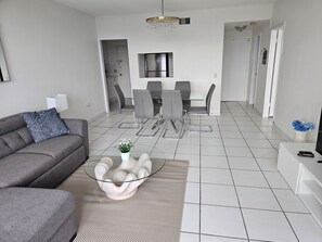 Ein modernes Wohnzimmer mit einem grauen Sofa, einem Glastisch und einem Essbereich mit grauen Stühlen und einem weißen Tisch.