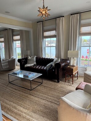 Ein Wohnzimmer mit einem Ledersofa, einem Glastisch, einer Lampe und Blick auf ein Gewässer durch große Fenster.