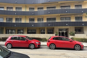 Ein mehrstöckiges Gebäude mit gelber Fassade, davor zwei rote Autos und ein Schild, das auf einen Wohnbereich hinweist.
