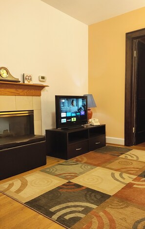 A living room with a flat-screen TV, a fireplace, a clock, and a patterned rug. - Cosy Retro 2 bedroom Cottage in vibrant Broad Ripple Village Indianapolis (Indianapolis)