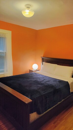 A room with an orange wall, a wooden bed with white pillows, a nightstand with a lamp, and a window with a curtain. - Cosy Retro 2 bedroom Cottage in vibrant Broad Ripple Village Indianapolis (Indianapolis)