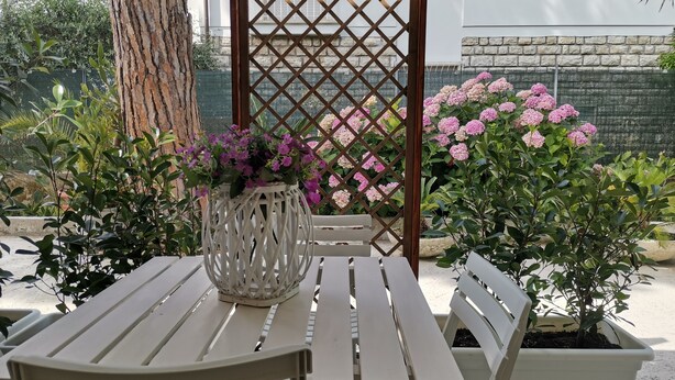 Un patio con mesa y sillas blancas, rodeado de macetas con plantas y una estructura de celosía.
