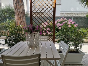 Eine Terrasse mit weißem Tisch und Stühlen, umgeben von Kübelpflanzen und einem Gitterwerk.