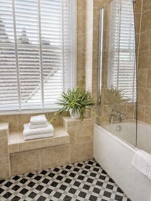 Ein Badezimmer mit Badewanne, Dusche, einem Fenster mit Jalousien und einem Blumentopf auf einem Regal.