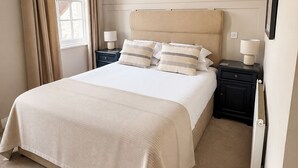 Un lit bien fait, avec une tête de lit beige, deux tables de chevet avec des lampes, une fenêtre avec des rideaux et un radiateur.