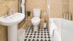 Une salle de bain comprenant un lavabo, des toilettes et une baignoire, avec un carrelage au sol et un miroir muni de lumières.