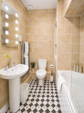 Ein Badezimmer mit Waschbecken, Toilette und Badewanne, gefliestem Boden und einem beleuchteten Spiegel.