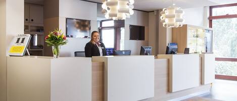 A hotel reception desk with a receptionist, a digital screen, and a large window.