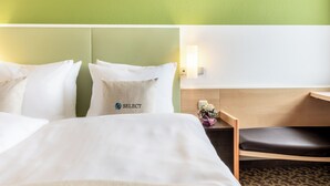 Habitación de hotel con cama, almohadas, mesita de noche, lámpara y una pequeña mesa con un jarrón de flores.