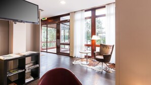 Habitación de hotel con televisión, estantería, sillón de cuero, mesita con lámpara y ventanas amplias con vistas a árboles.