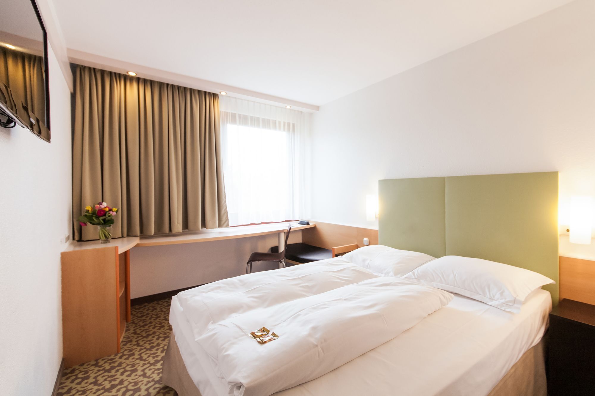 Une chambre d’hôtel avec un lit, un bureau, une chaise, un miroir et une fenêtre avec des rideaux.
