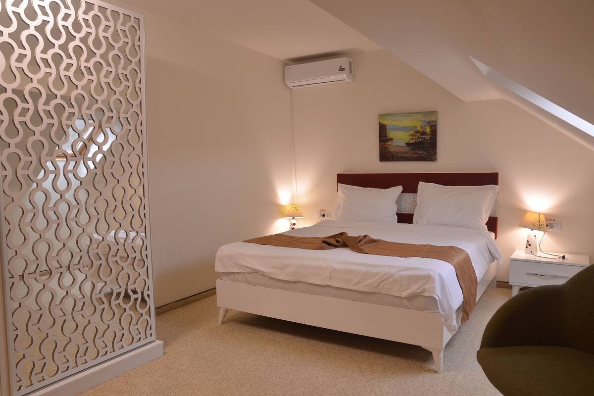 Un dormitorio con cama, lámparas de noche, un cuadro, aire acondicionado y un separador decorativo.