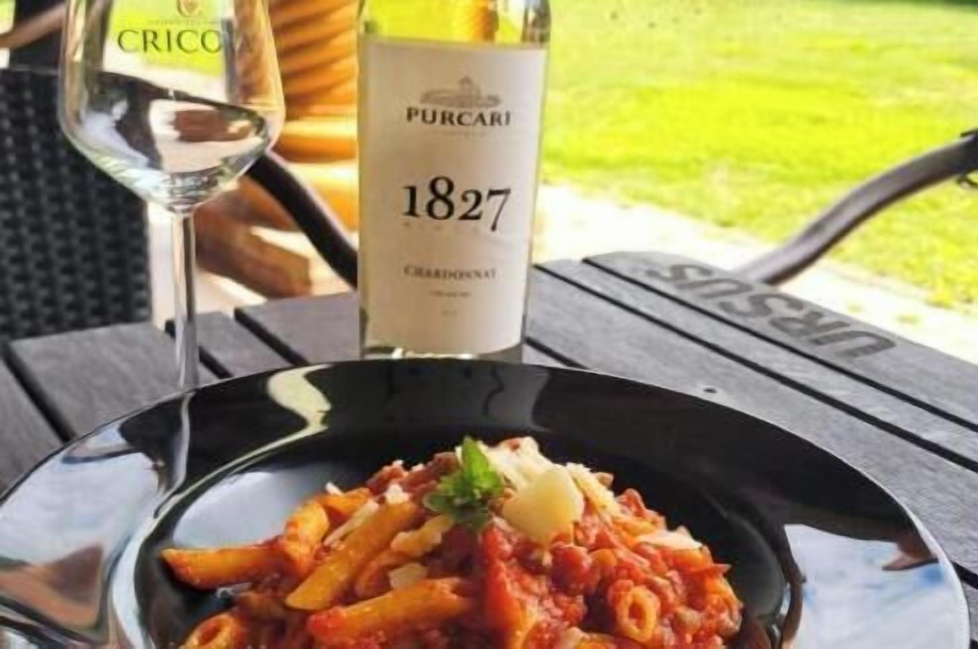 A bottle of Pucari 1827 Chardonnay and a plate of pasta with a glass of wine on a table.