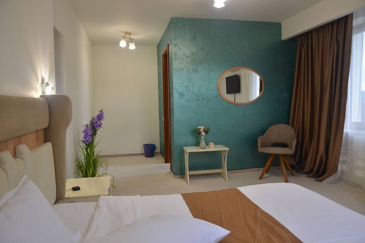 Una habitación de hotel con cama, una mesita con un jarrón, una silla, un espejo redondo y una pared de acento color aguamarina.