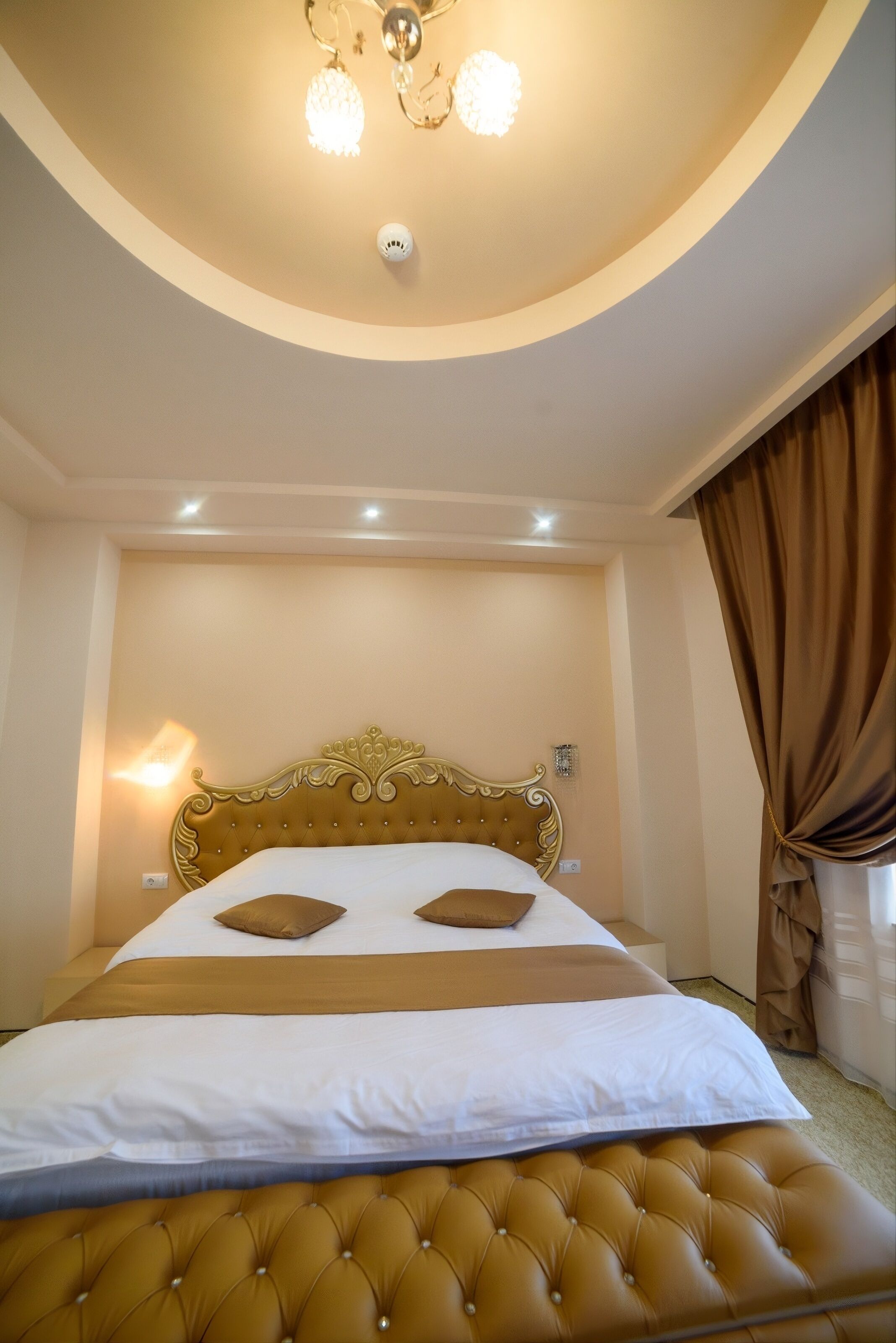 A hotel room with a tufted headboard, a bed with white and brown bedding, a brown upholstered bench, a wall-mounted light, and a ceiling light fixture.