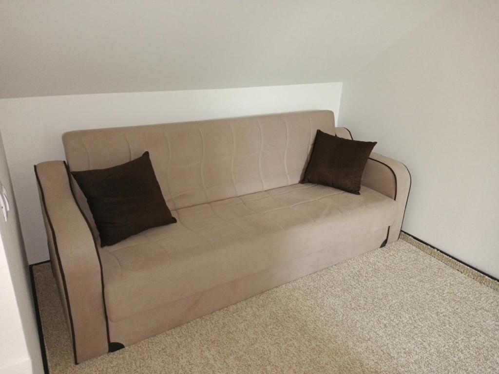 A beige sofa with two dark cushions in a room with a plain white wall and a carpeted floor.