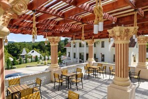 An outdoor patio with wooden tables and chairs, columns with decorative capitals, and a view of a white building with a red roof. - Virginia Gardens  (Baile Govora)