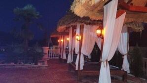 A covered outdoor seating area with thatched roofs and white drapes, illuminated by lantern-style lights. - Virginia Gardens  (Baile Govora)
