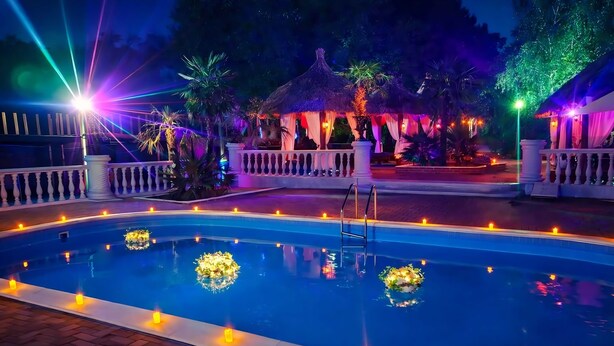 A swimming pool with candles and flowers, surrounded by a white fence and palm trees. - Virginia Gardens  (Baile Govora)