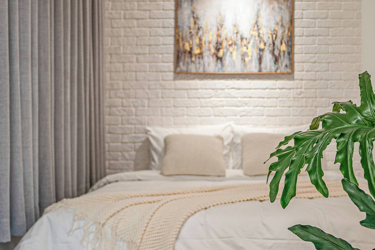 Un dormitorio con pared de ladrillo blanco, cama con ropa de cama blanca, un cuadro abstracto grande y una planta verde.
