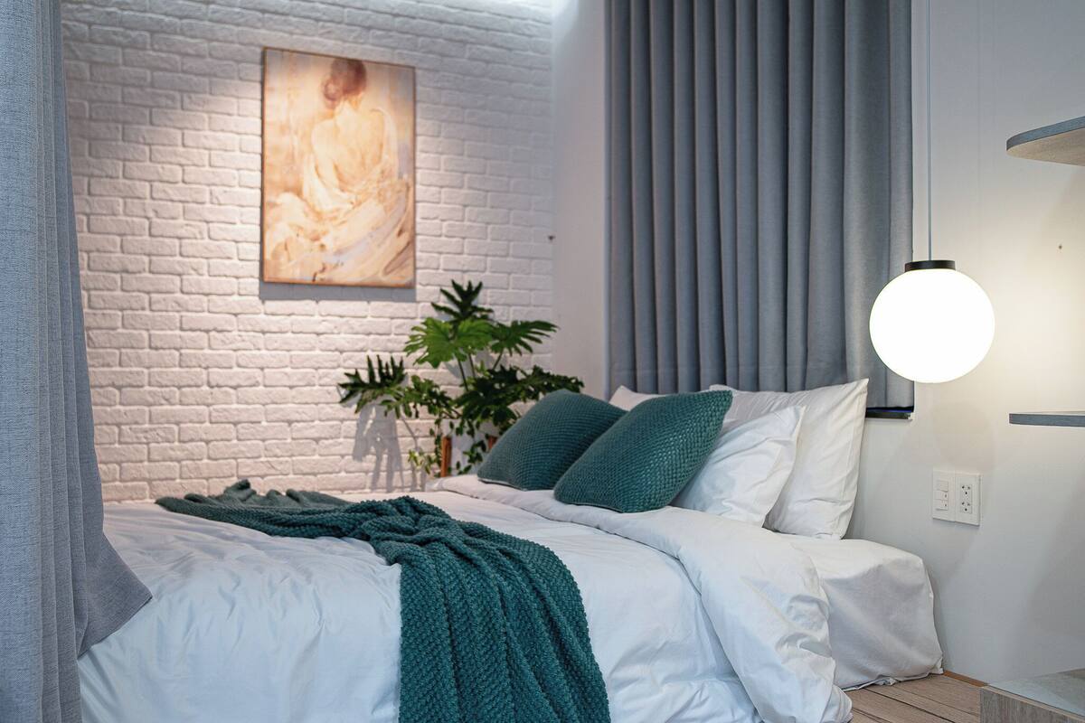 Un dormitorio moderno con una pared de ladrillo blanco, un cuadro, una planta verde y una cama con ropa de cama blanca y almohadas color turquesa.