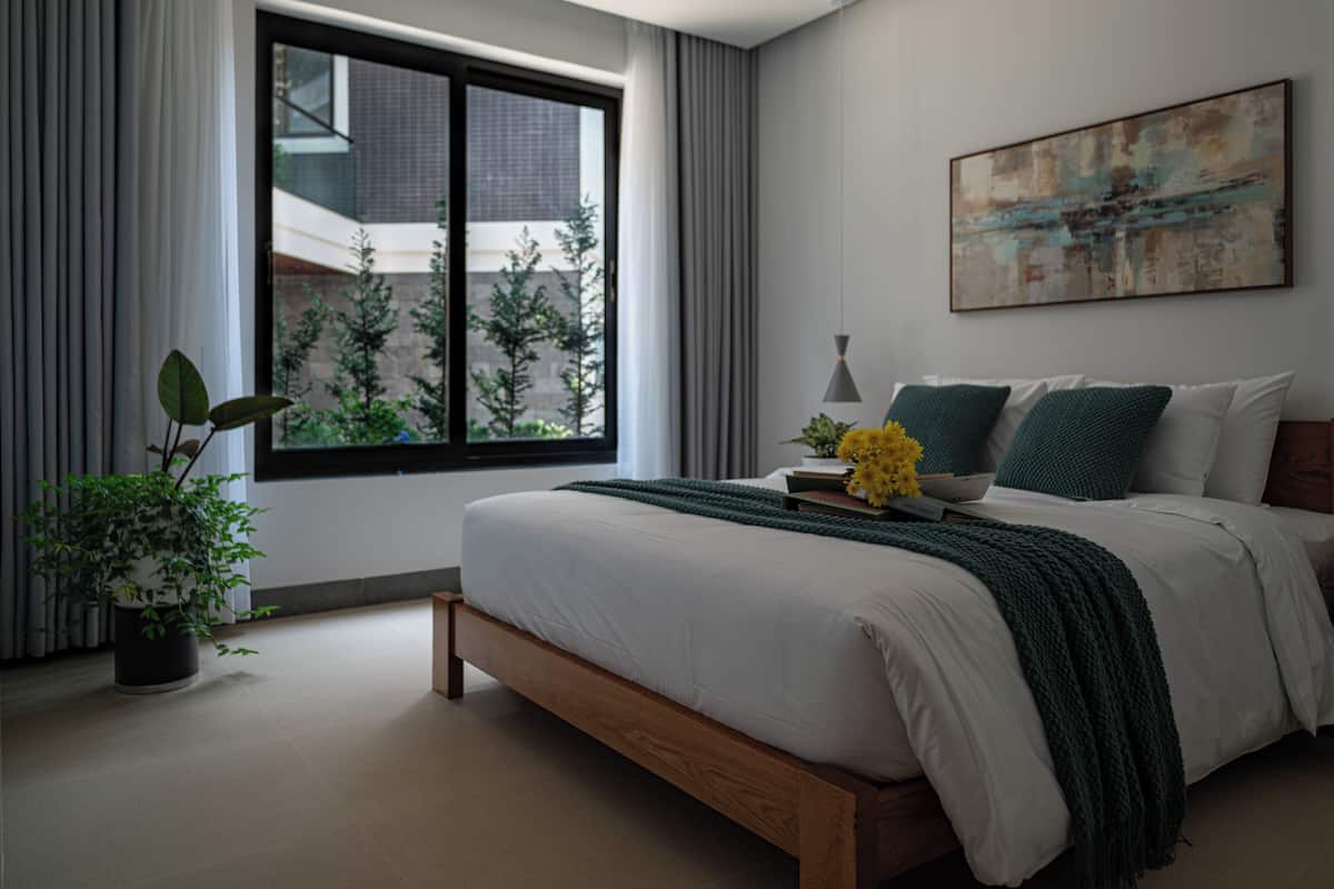 Un dormitorio moderno con un ventanal grande, una cama con estructura de madera, una mesita de noche con lámpara y una planta en maceta.