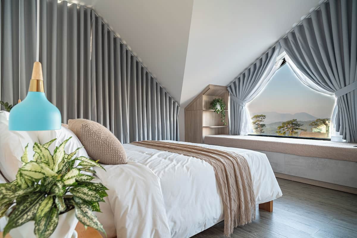 Un dormitorio con cama, ventana con cortinas, una planta y vistas al exterior.