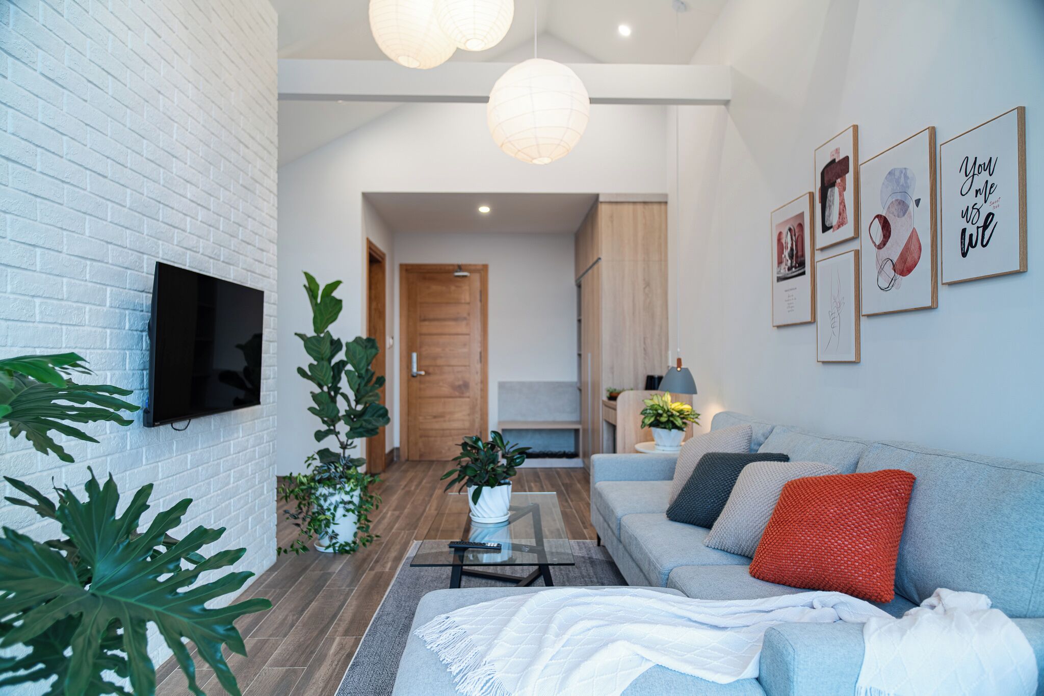 Una sala de estar moderna con un sofá, una mesa de centro y un televisor de pantalla plana montado en una pared de ladrillo blanco. Hay cuadros enmarcados en la pared y una puerta de madera al fondo.