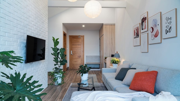 A modern living room with a sofa, a coffee table, and a flat-screen TV mounted on a white brick wall. There are framed pictures on the wall and a wooden door in the background. - Zenora Hillside Garden (Xuan Huong - Da Lat)
