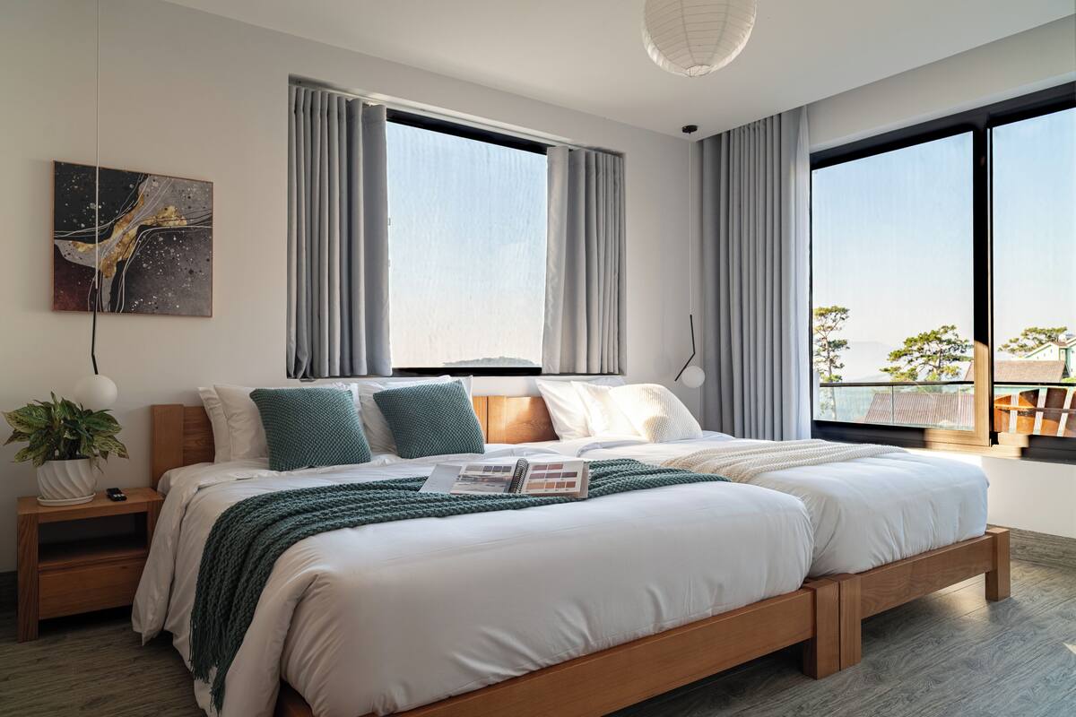 Un dormitorio moderno con una cama grande, dos ventanas, vistas al exterior, una mesita de noche con lámpara y una planta.