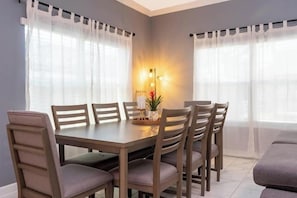 A dining area with a wooden table and six chairs, two windows with sheer curtains, and a floor lamp with three lights. - SL2964PS - Luxurious 5Bdrooms Villa with Priv Pool (Kissimmee)