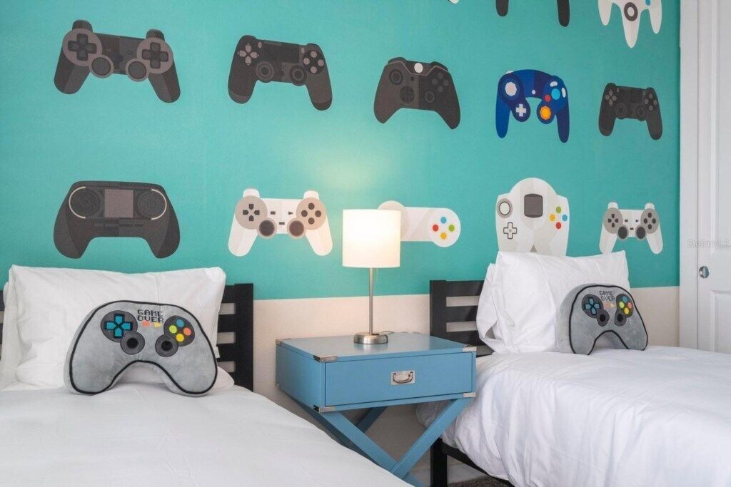 A bedroom with a teal wall decorated with various game controller designs, a bedside table with a lamp, and two beds with pillows featuring game controller designs.