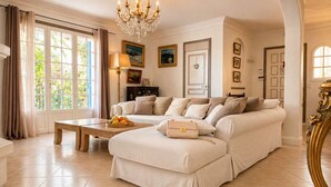 A spacious living room with a large sofa, a coffee table with fruit, a chandelier, and a view of greenery outside. -  Super Cannes villa D Aurus (Vallauris)
