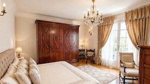 A bedroom with a large bed, a wooden wardrobe, a chandelier, and a sitting area with a chair and table. -  Super Cannes villa D Aurus (Vallauris)