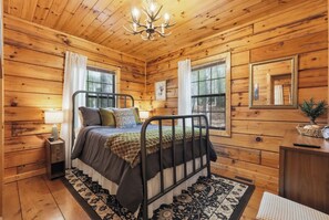 Quarto de cabana de madeira com uma cama, mesas de cabeceira, um candelabro e uma janela com vista para árvores.