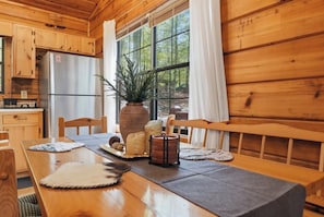 Interior de uma cabana de madeira com mesa de jantar, cadeiras, geladeira, janela grande com cortinas e um vaso de planta.